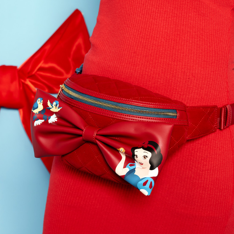 Close up image of a woman in a red dress standing against a blue background wearing the Loungefly Snow White Classic Apple Belt Bag around her waist, featuring a 3D bow featuring Snow White on one side with a bird and two birds on the other side of the bow
